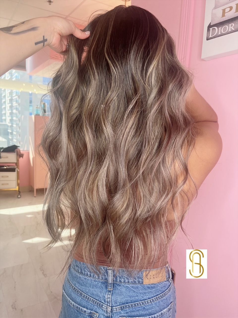 A woman with long wavy hair is standing in front of a pink wall.