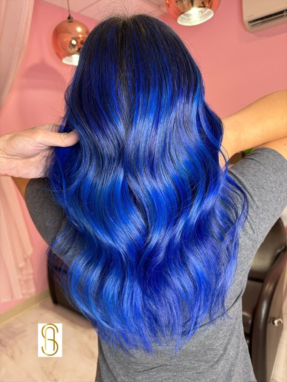 A woman with long blue hair is standing in front of a pink wall.