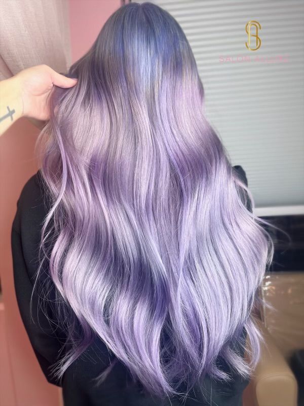 A woman with long purple hair is standing in front of a window.