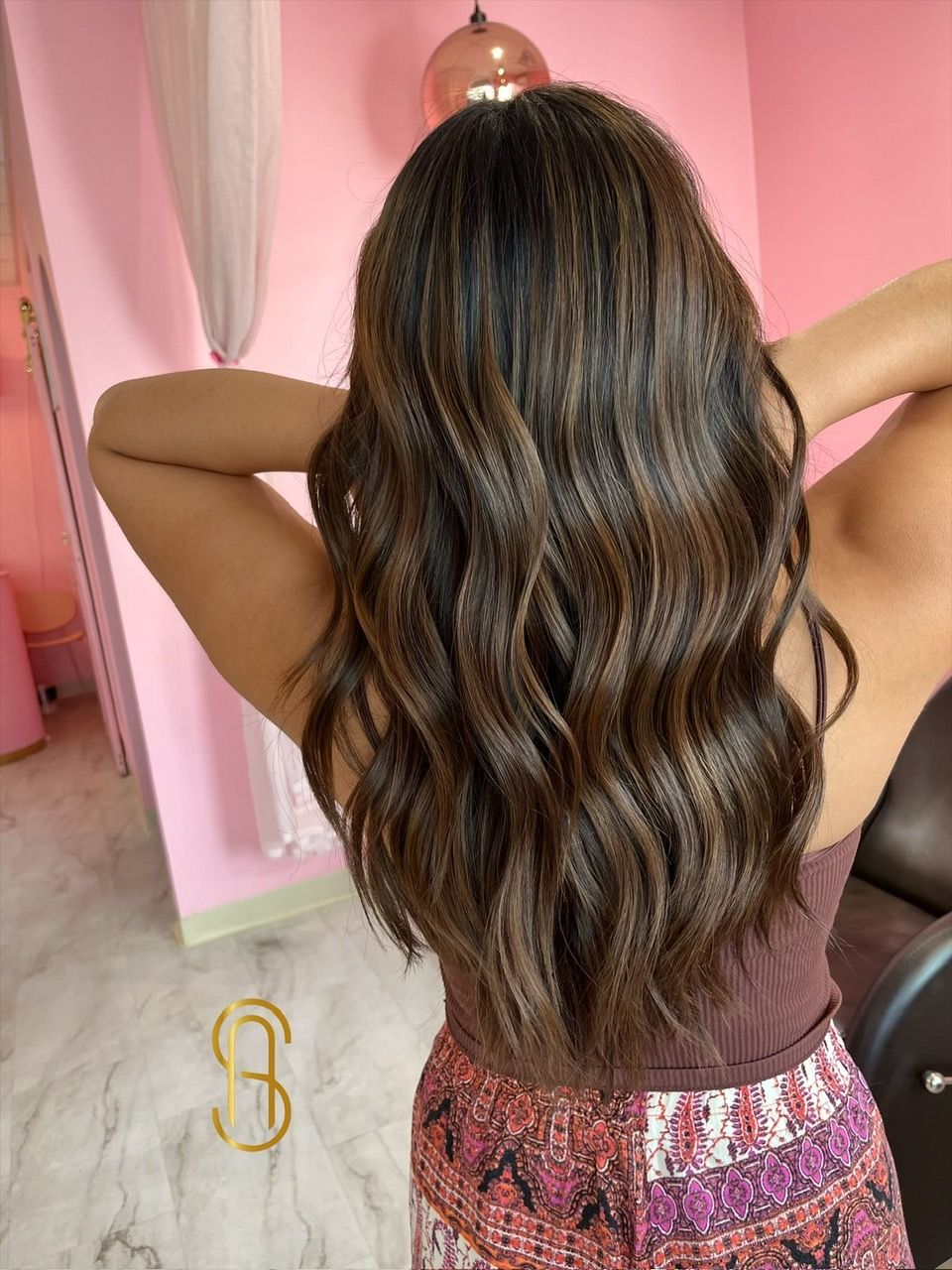 A woman with long brown hair is standing in a room with her hands behind her head.