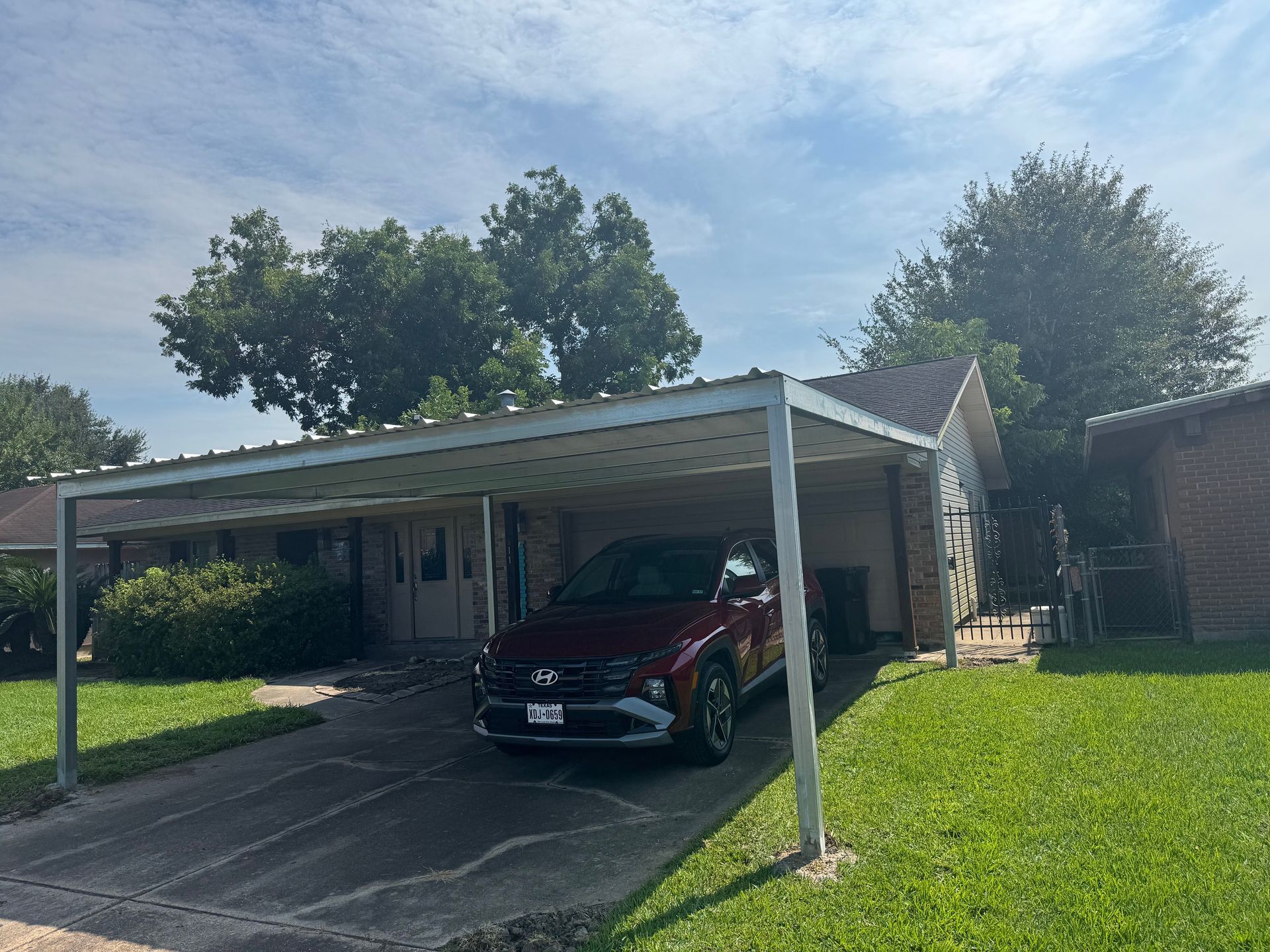 Red car parked under a carport in front of a house with trees and grass on a sunny day.