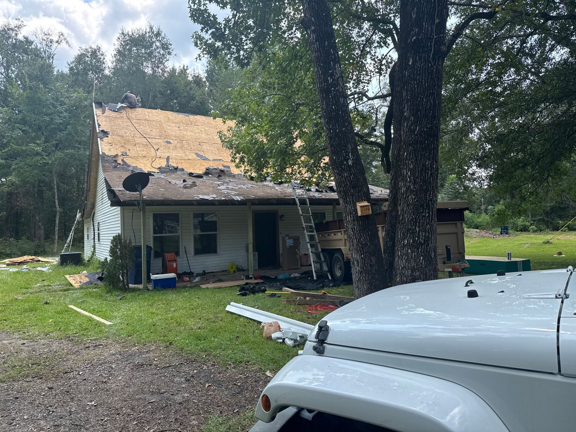 House with roof damage, tools, and a white Jeep. Outdoors, overcast sky, forest in background.
