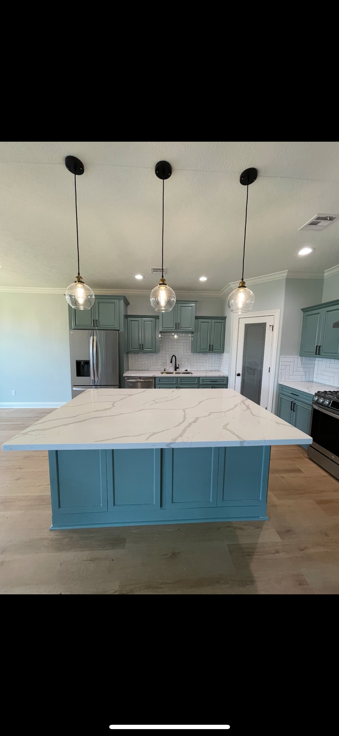 A teal kitchen island with a white countertop under three pendant lights. Kitchen cabinets and appliances visible.