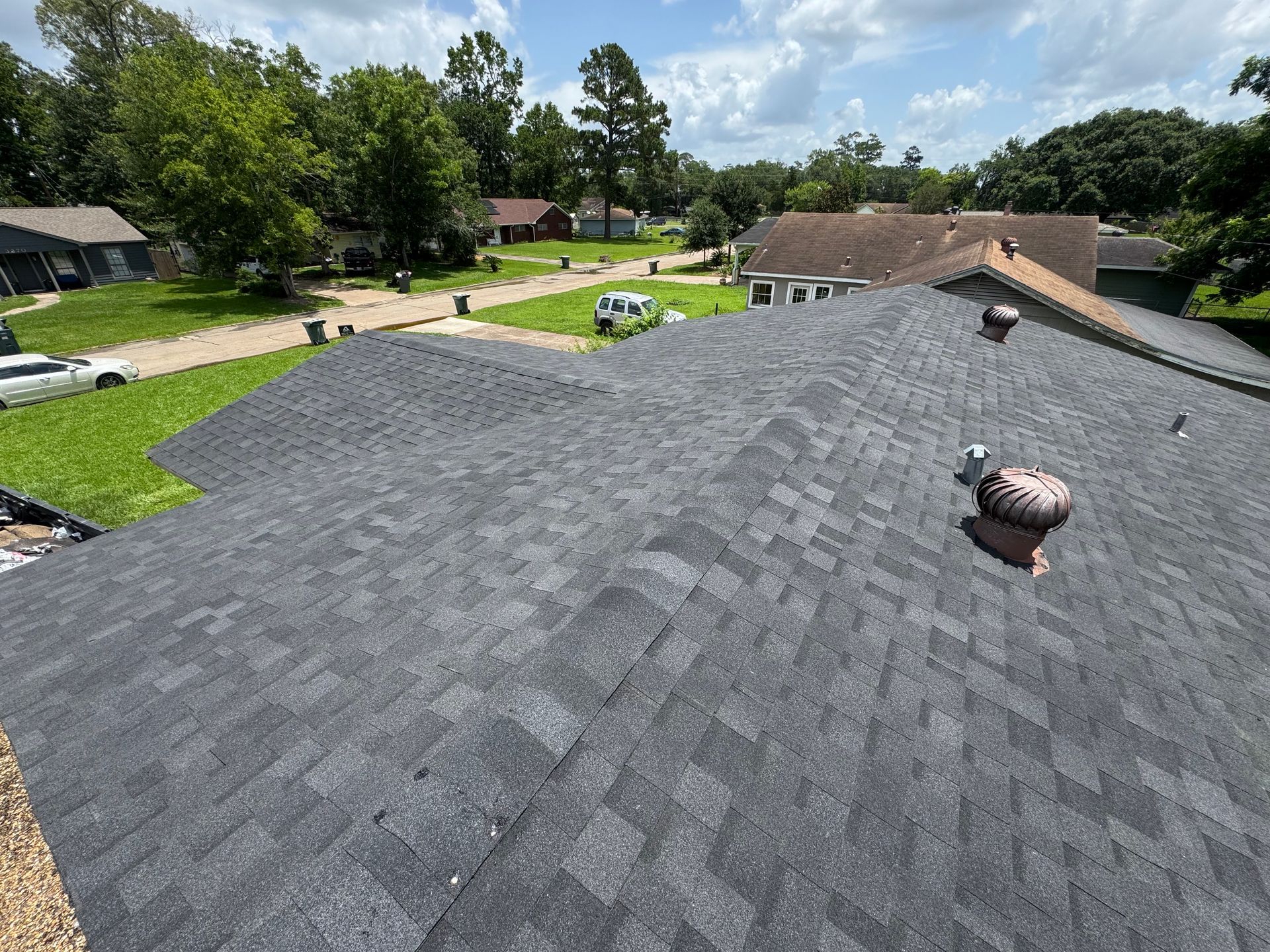 Gray asphalt shingle roof of a house with vents; green yard and houses in the distance.