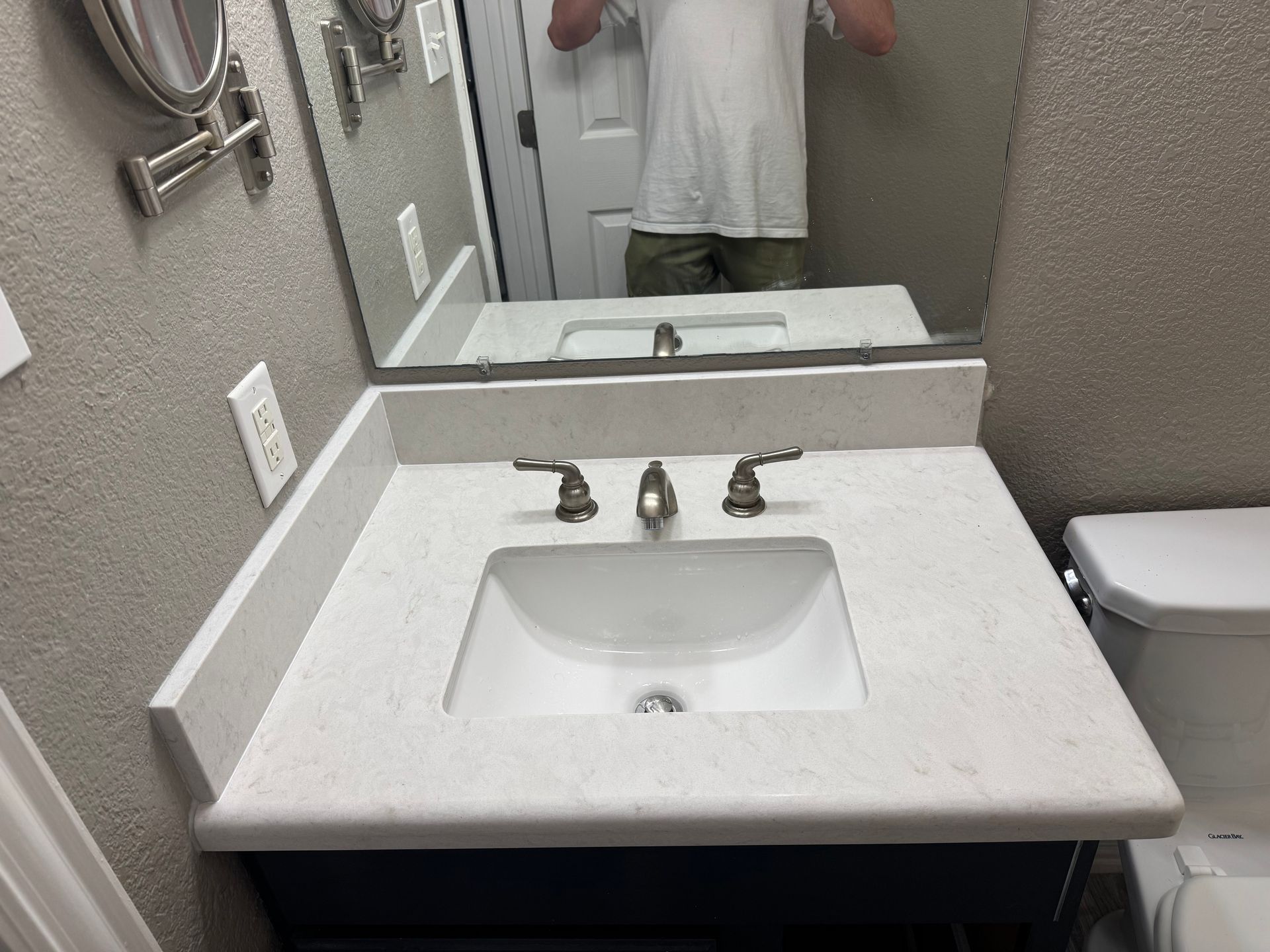 Bathroom vanity with white countertop, sink, and mirror. A person is reflected in the mirror.