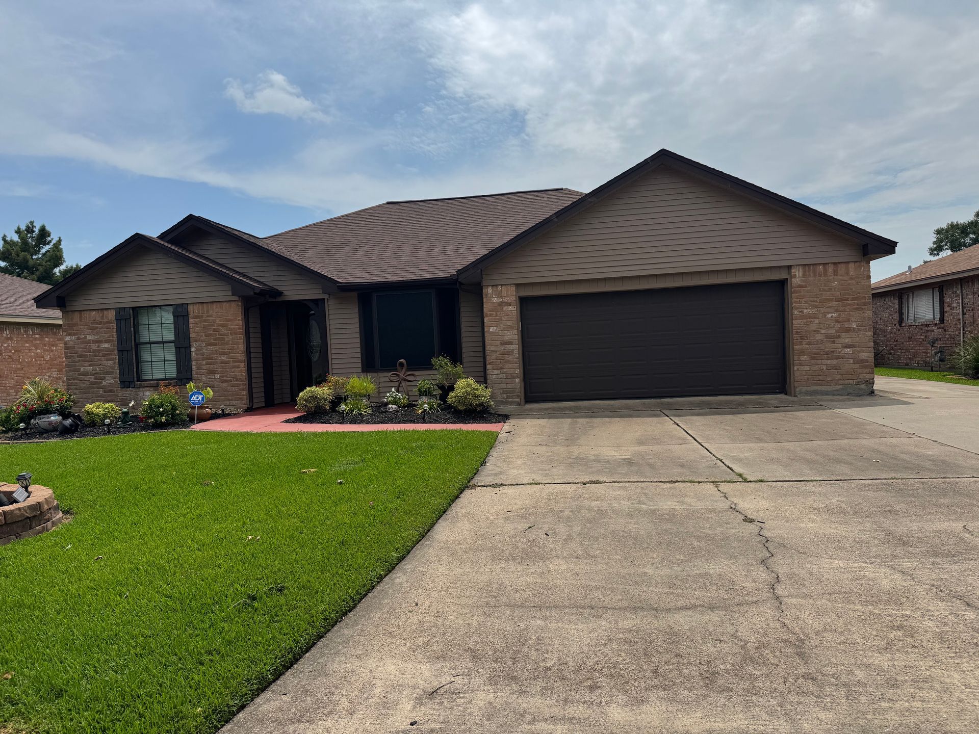 Brick house with brown roof and garage door, green lawn, and concrete driveway on a sunny day.