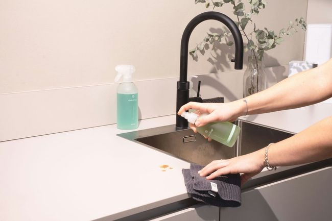 Hands washing a cloth in a kitchen sink beside a green spray bottle.