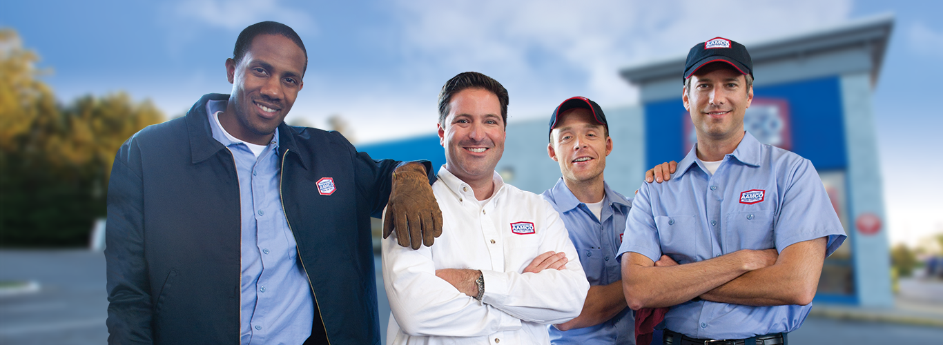 Four people standing in front of a building, smiling. They wear AAMCO work uniforms. The sky is blue.