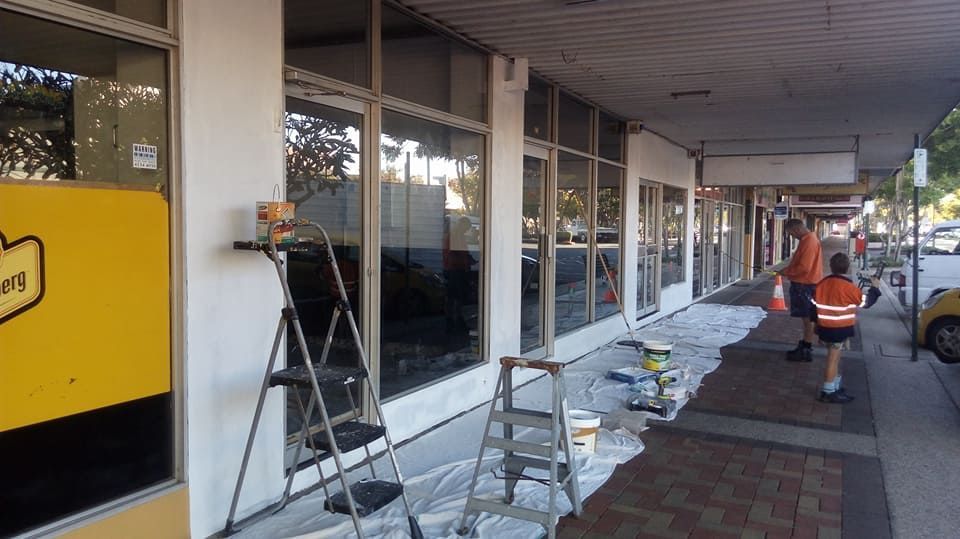 A Man Is Walking Down a Sidewalk Next to A Building that Is Being Painted — Your Local Handywoman in Burnett Heads, QLD