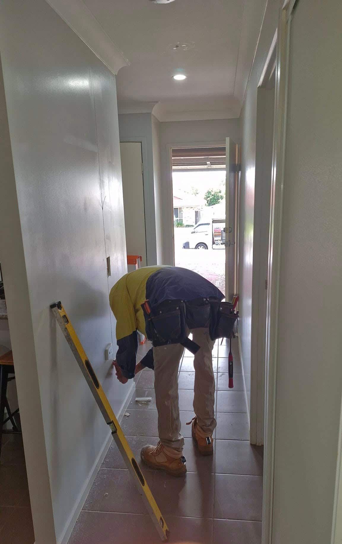 A Man Is Kneeling Down in A Hallway with A Tape Measure — Your Local Handywoman in Burnett Heads, QLD