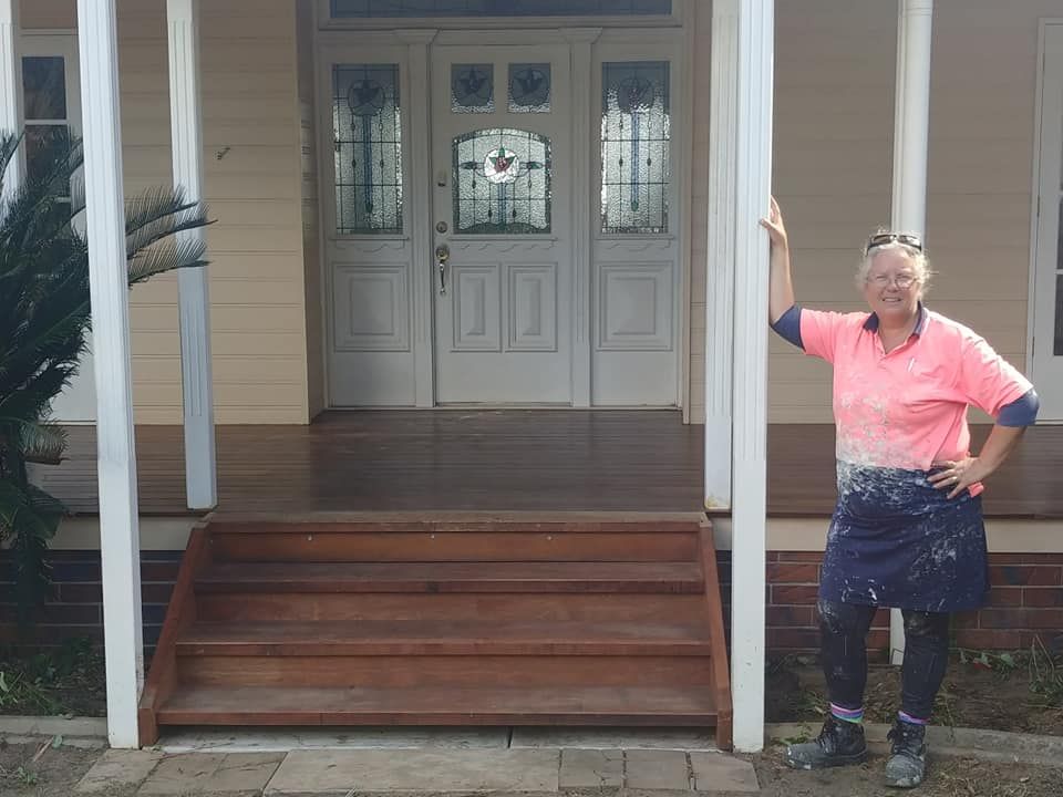 A Woman in A Pink Shirt Is Standing in Front of A House — Your Local Handywoman in Burnett Heads, QLD