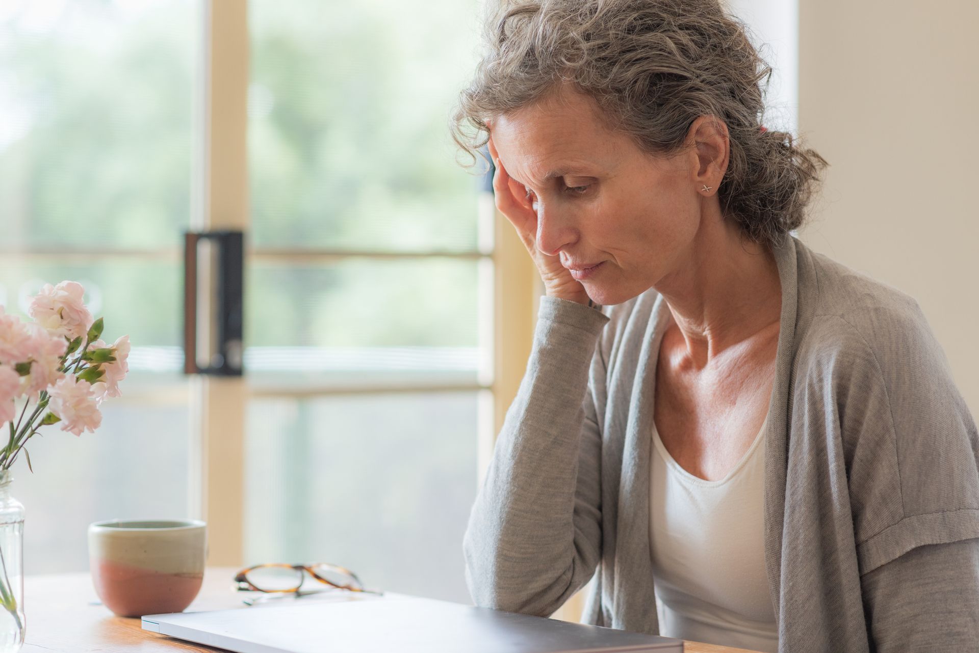 Woman with grey hair resting head on hand, looking down, laptop and coffee cup on table.