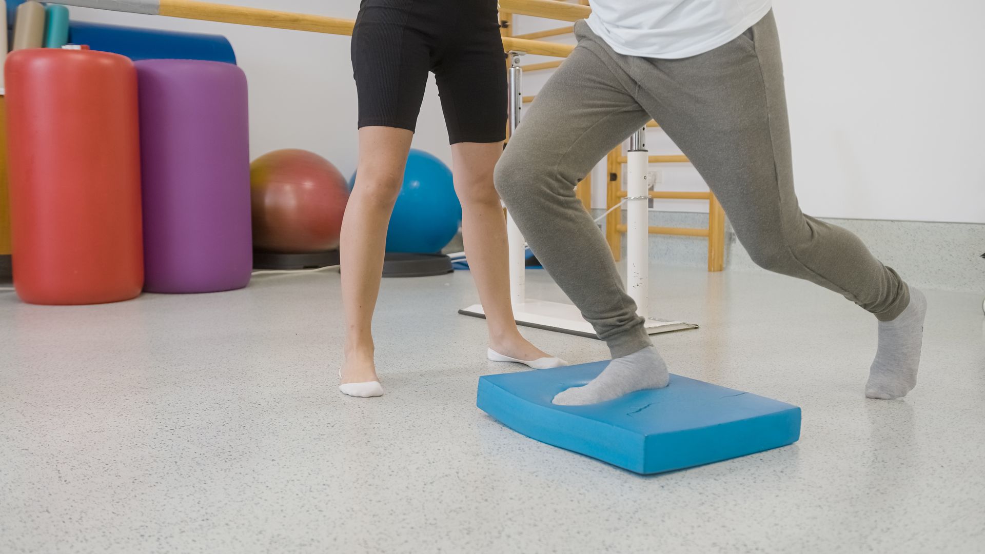 Man performing a step-down exercise on a blue foam block, supervised by a therapist in a physiotherapy room.