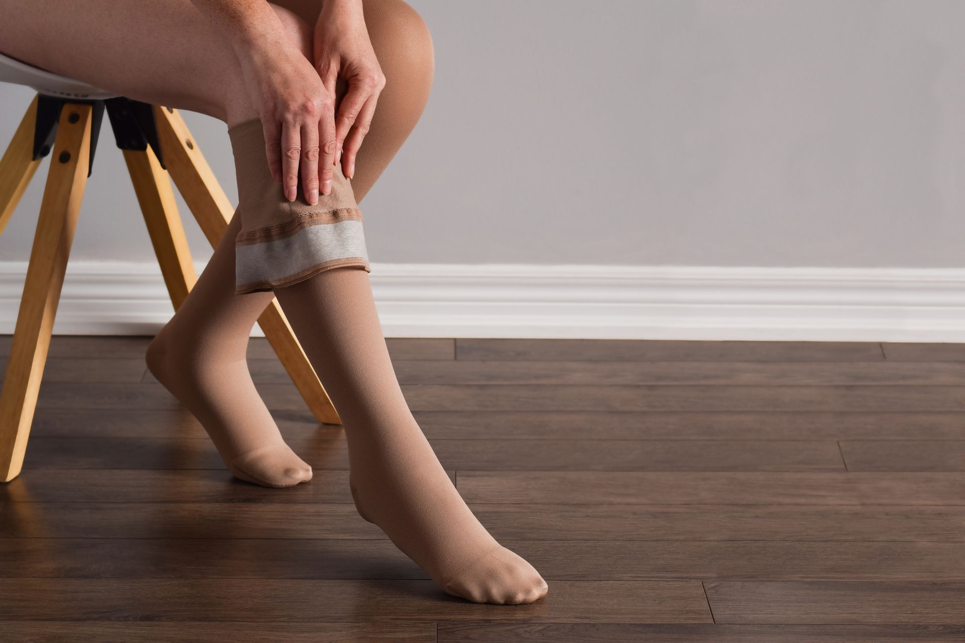 Person seated, adjusting compression stocking on leg; neutral background, wooden floor.