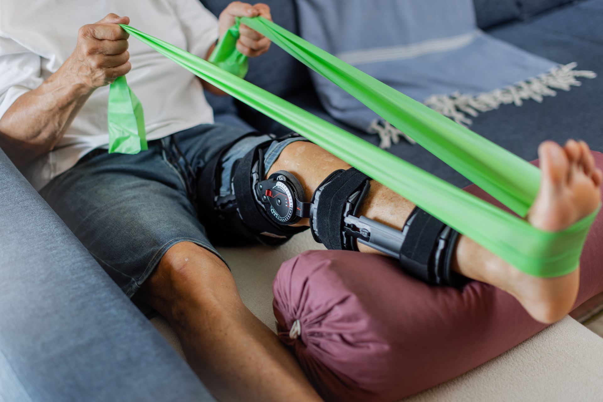 Person with a knee brace doing physical therapy with a green resistance band, in a home setting.