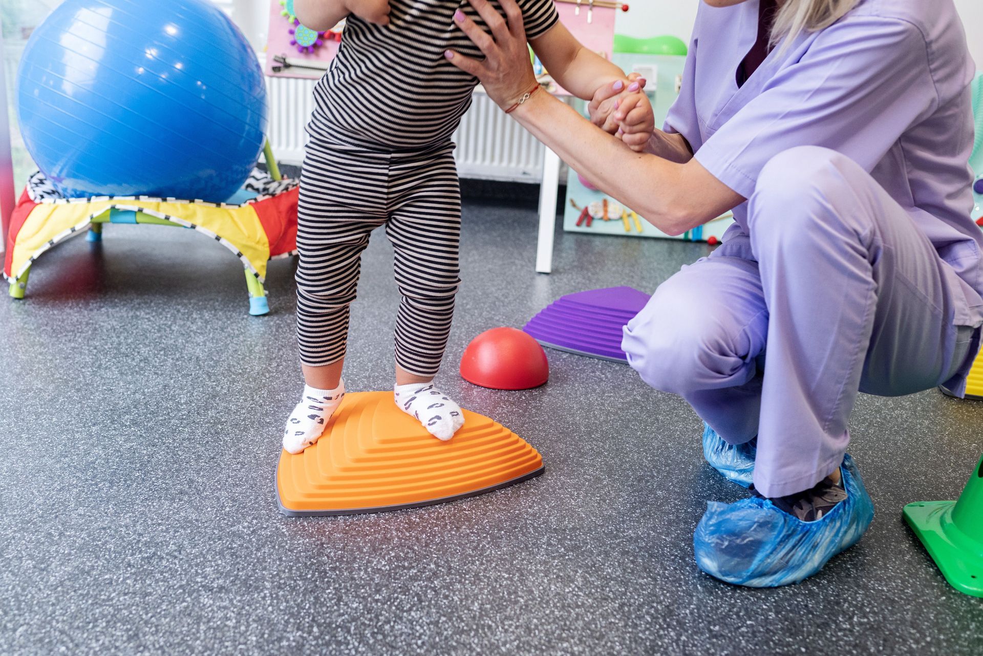 A child in striped pants walks on an orange balance step, assisted by a person in scrubs. Gym setting.