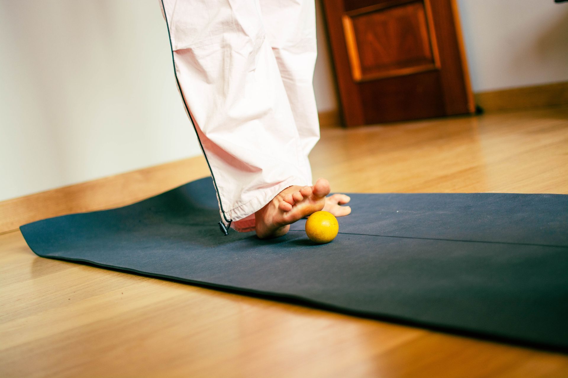 Foot rolling a yellow ball on a black exercise mat, indoors. Pink pants, hardwood floor.