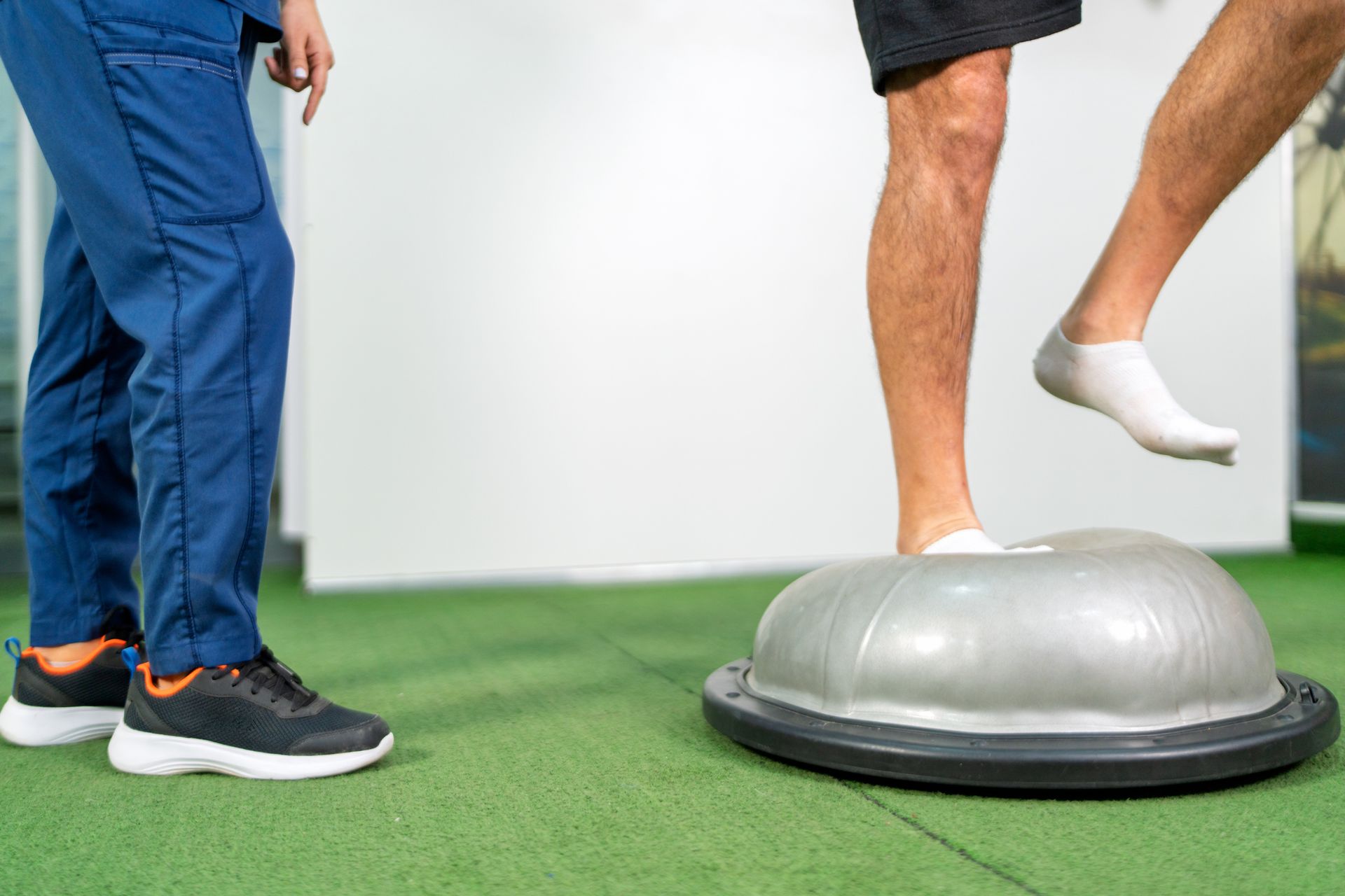 Person balancing on a Bosu ball with assistance from a medical professional. They are in a clinic setting.