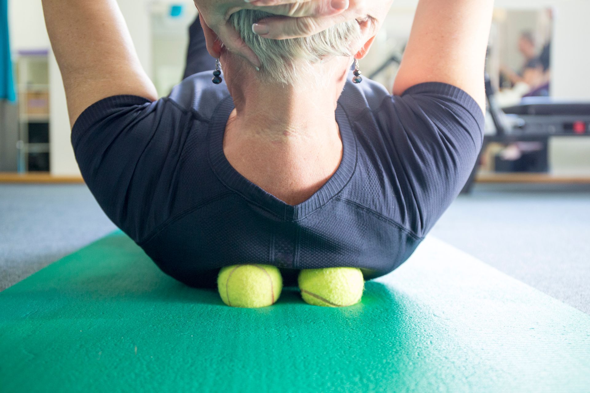 Person lying on back, using tennis balls to massage upper back on a green mat.