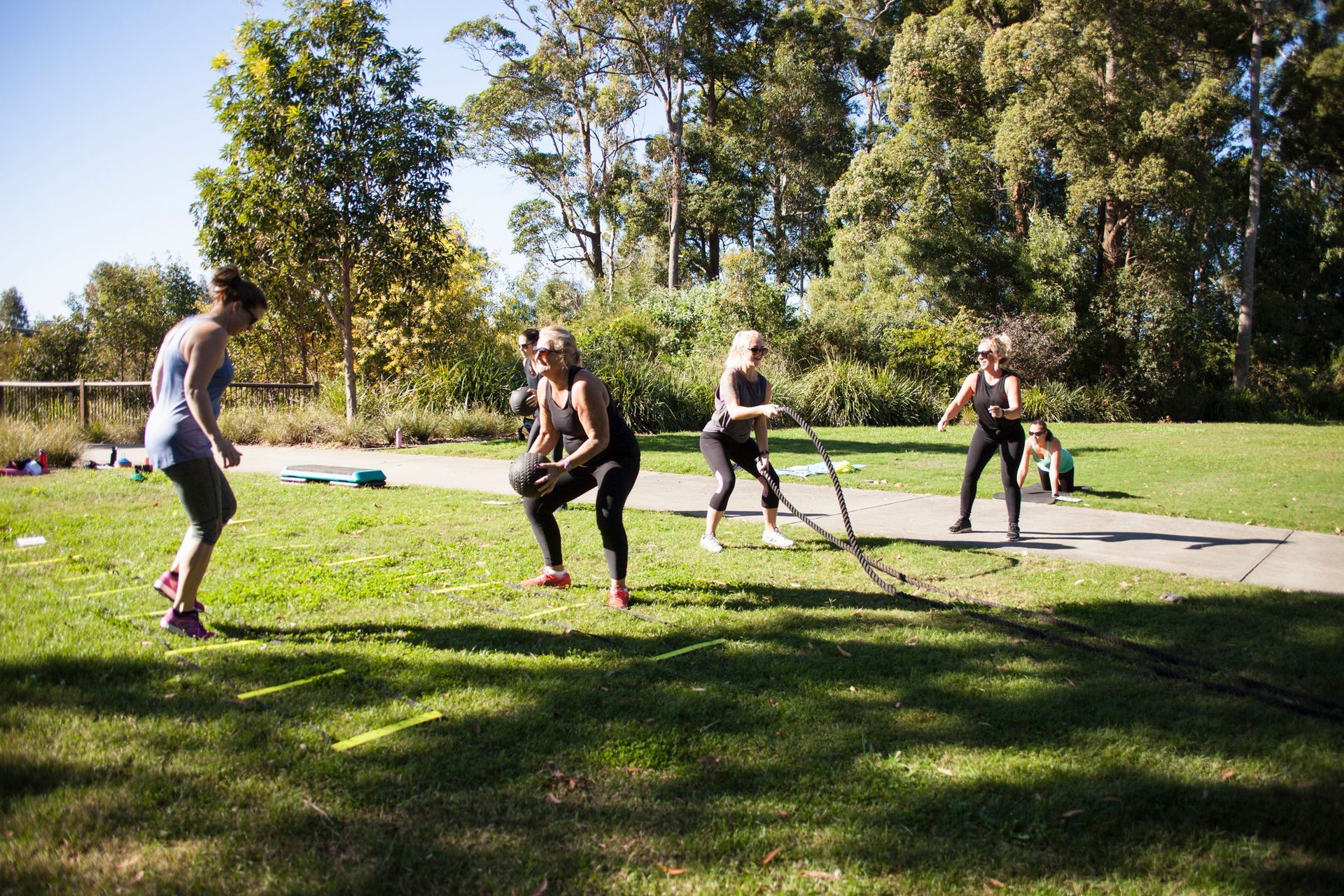 Group of people exercising outdoors on a grassy area, using battle ropes and other equipment, under a sunny sky.