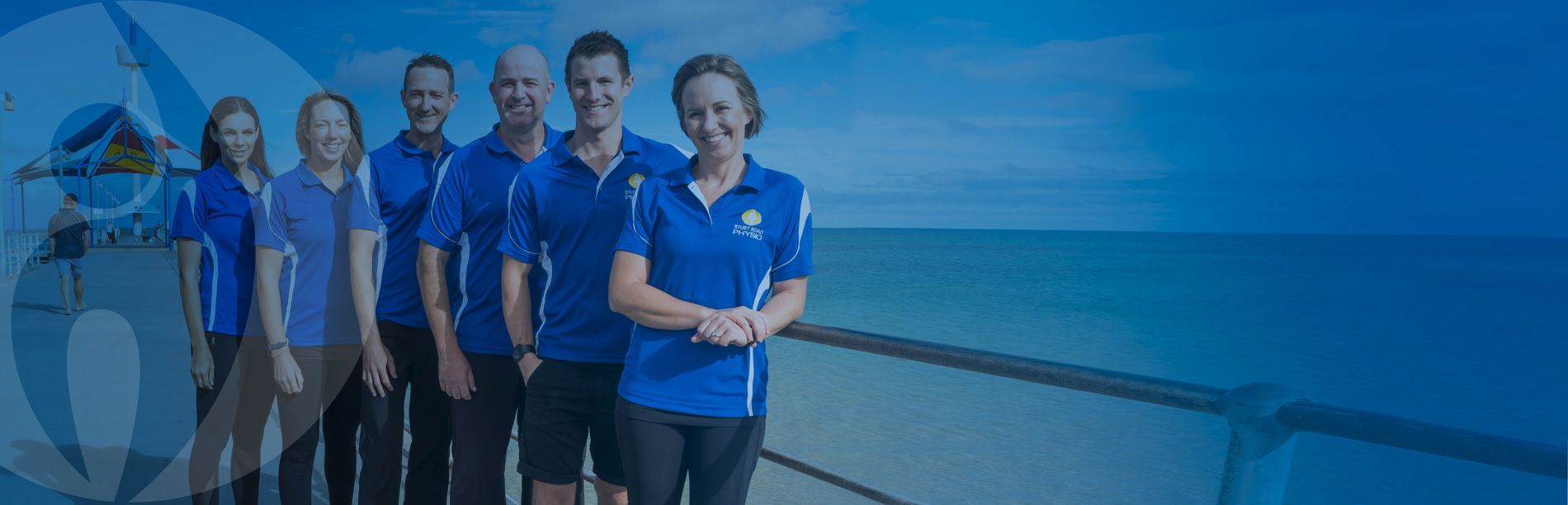 Six people in blue shirts standing on a pier overlooking the ocean.