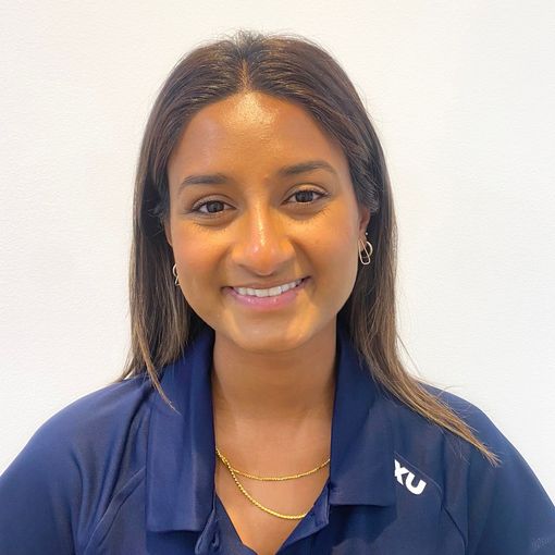 Woman smiling, wearing a blue shirt with gold necklace, in front of a white background.
