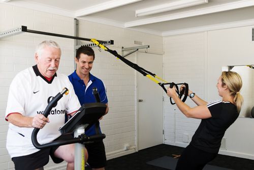 People in gym: Man on bike, another watching, woman using TRX. Bright, indoor setting.