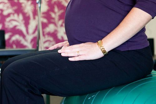 Pregnant person sits on a green exercise ball, hands resting on thighs, wearing black pants and purple top.