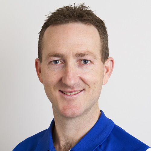 Man with short brown hair, smiling, wearing a blue polo shirt, against a white background.