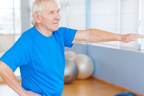 Older person in a blue shirt stretching arm out in a light-filled room with exercise balls in the background.