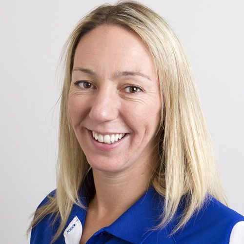 Blonde woman in blue shirt smiles, facing the camera against a neutral background.