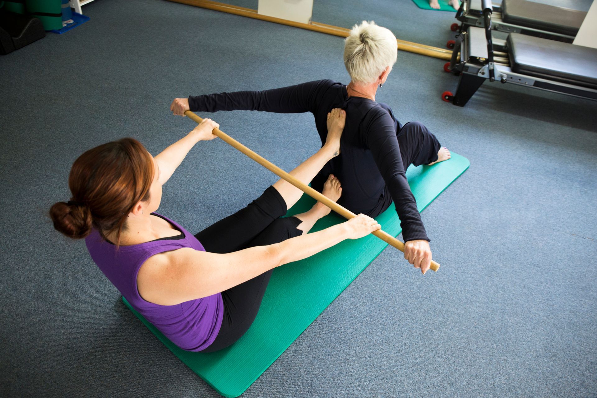 Woman assists another with a Pilates exercise using a wooden dowel on a mat.