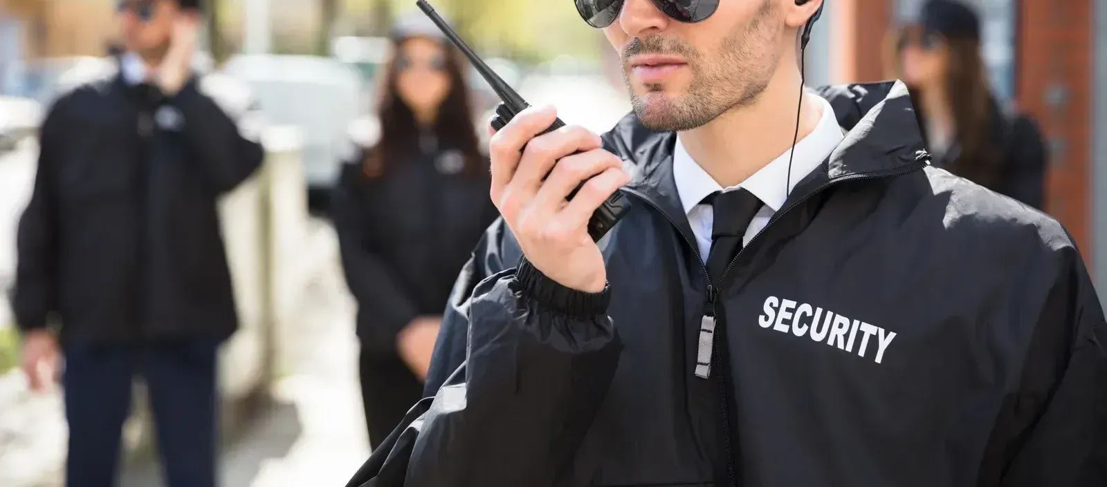 Security guard holding a radio, speaking. Other guards behind him. Outdoors, daytime.