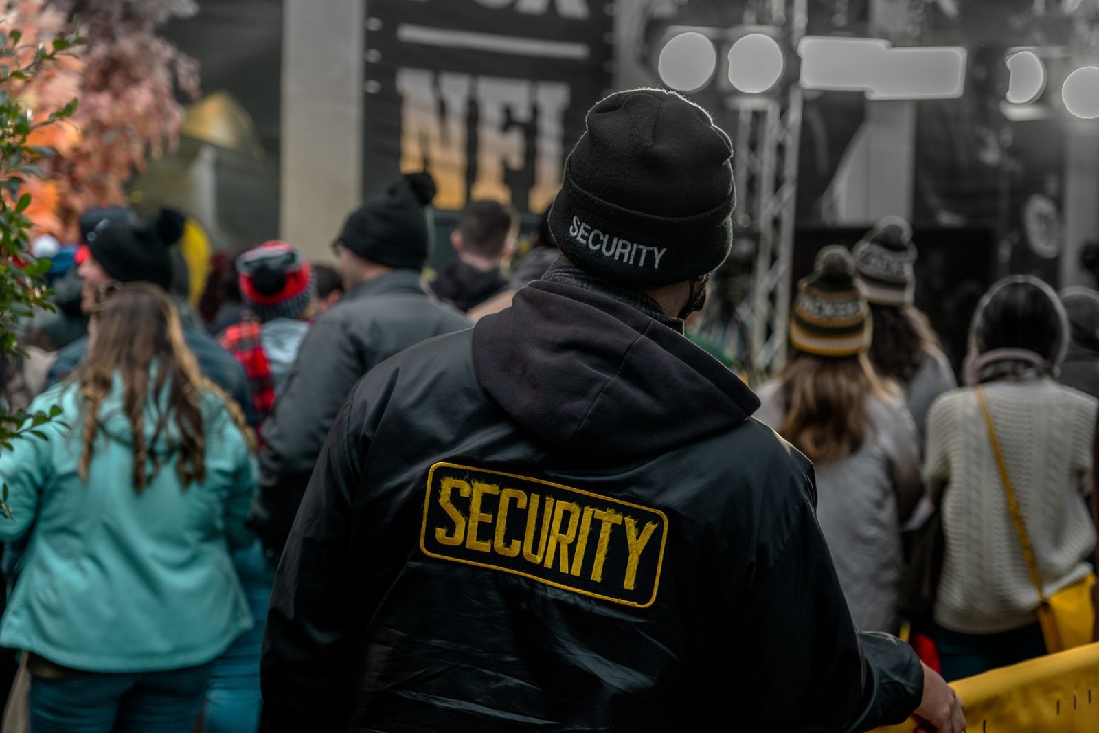 Security guard in black jacket and beanie stands before a crowd at an outdoor event.