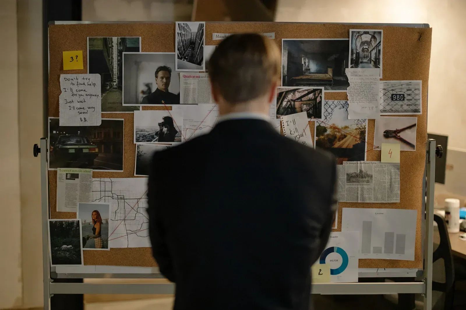 Man in a suit facing a corkboard covered with photos, notes, and charts.