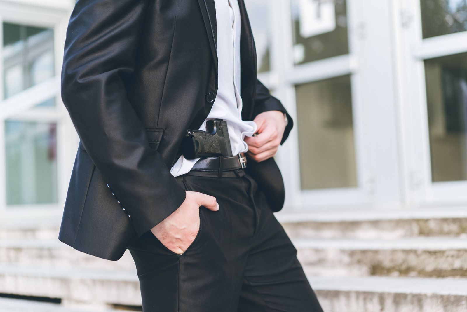 Man in black suit, hand on concealed handgun at waist. Outside a building.