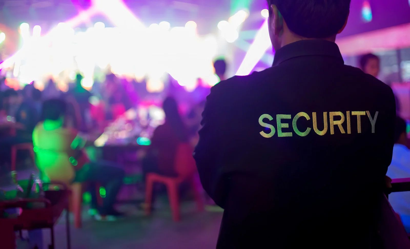 Security guard in black uniform watches over a crowded nightclub with colorful lights.