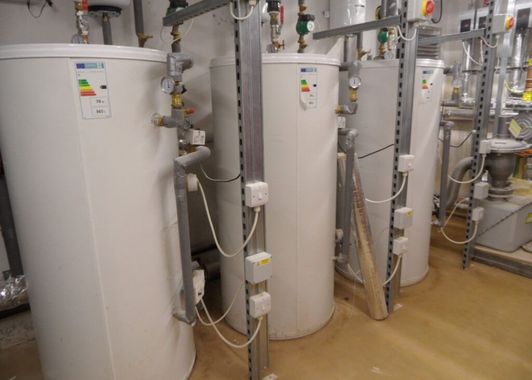 Four white water heaters with piping and electrical boxes in a utility room.