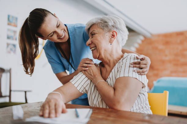 A smiling nurse in blue scrubs comforts an elderly woman, both with arms around each other, inside a building.