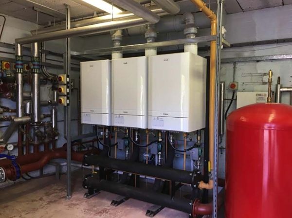 Three white boilers in a utility room with pipes, a red tank, and industrial setting.