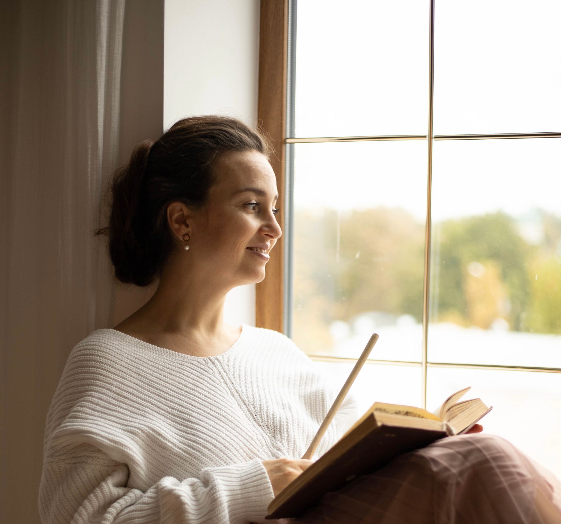 Женщина сидит на подоконнике и читает книгу.