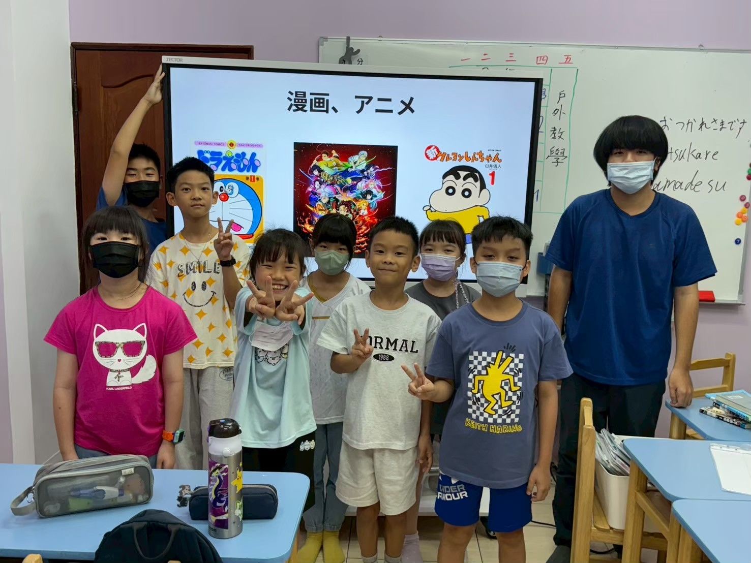一群孩子和一位老師在教室裡，螢幕上播放著日本動漫。