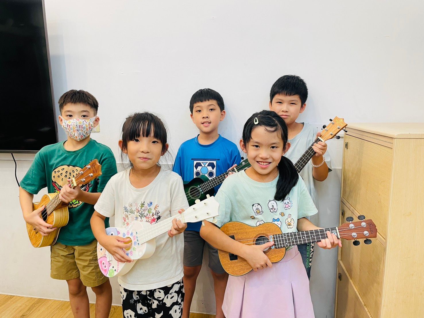 五個孩子在燈光明亮的房間裡抱著烏克麗麗琴。有些孩子對著鏡頭微笑。