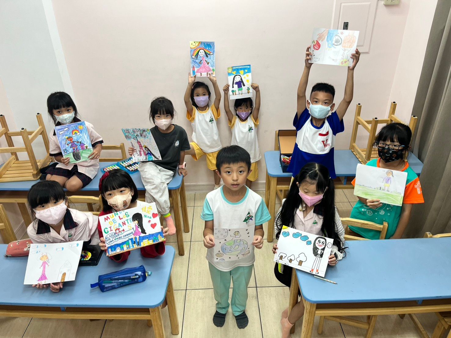 孩子們舉著畫作，可能是課堂上的作業。有些孩子戴著口罩，坐在課桌前。
