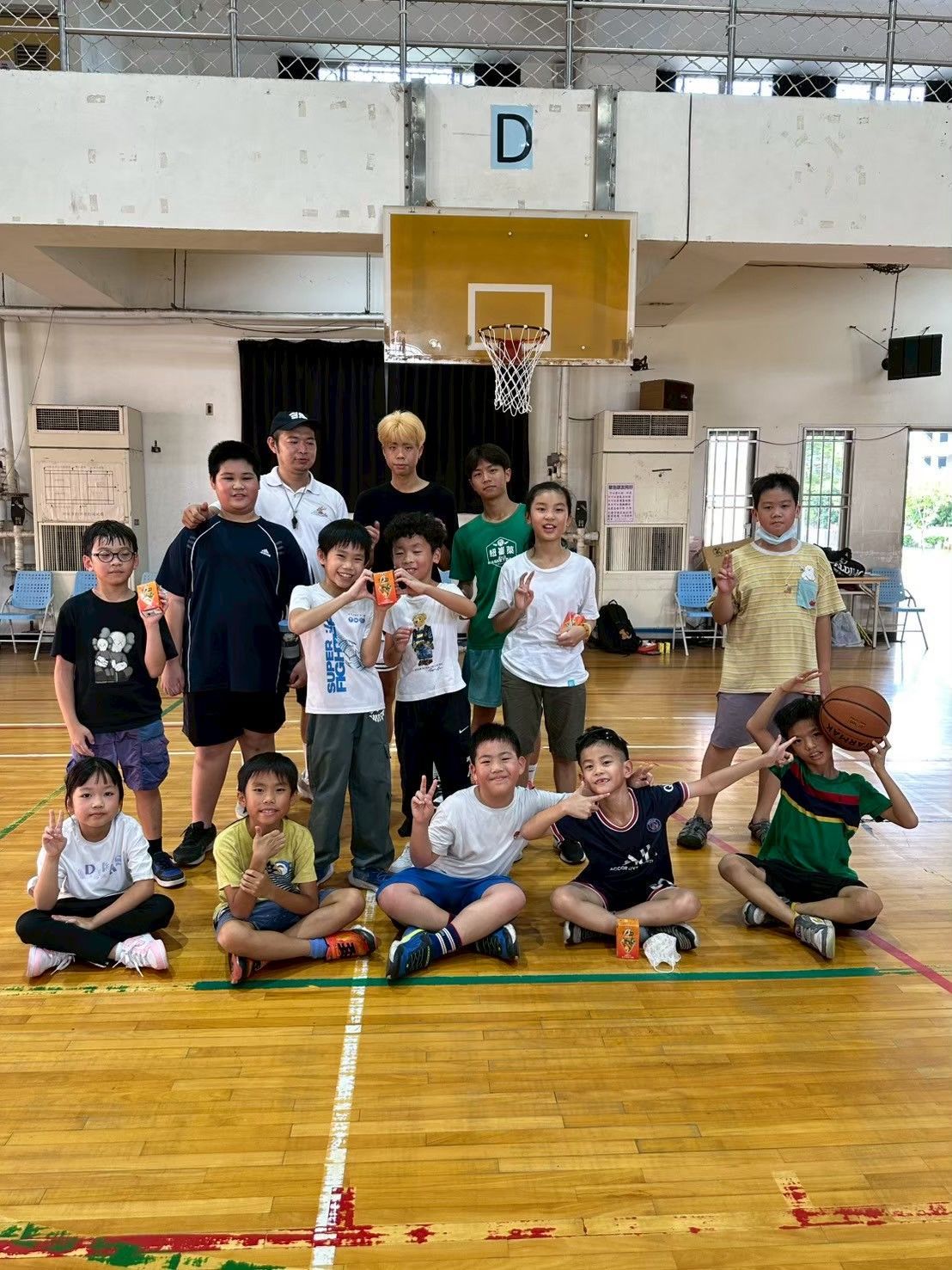 一群小孩和大人在籃球場上擺姿勢拍照。有些人拿著籃球；有些人擺出和平手勢。