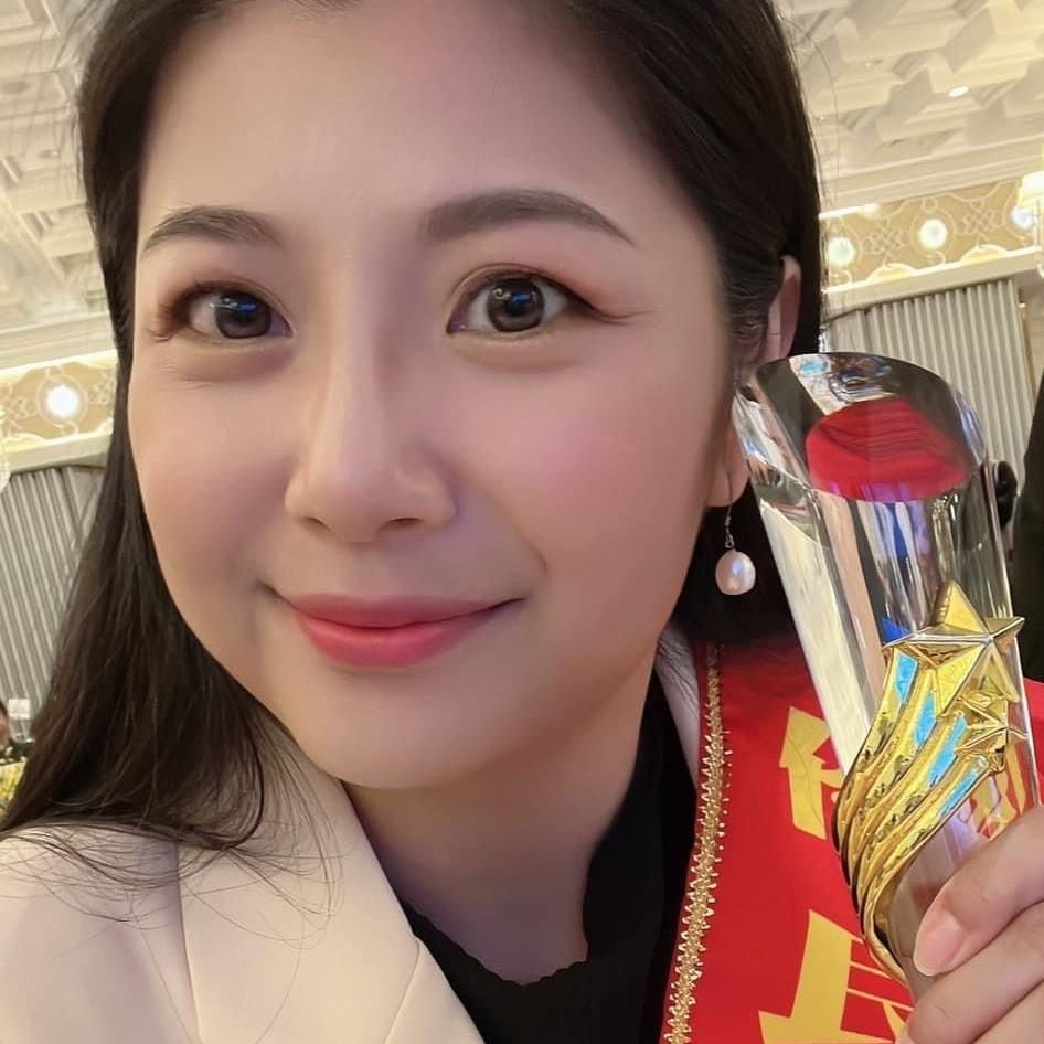 一位面帶微笑的女子手捧獎杯,獎杯上繫著紅色瑤帶。室內光線充足,天花板裝潢華麗。