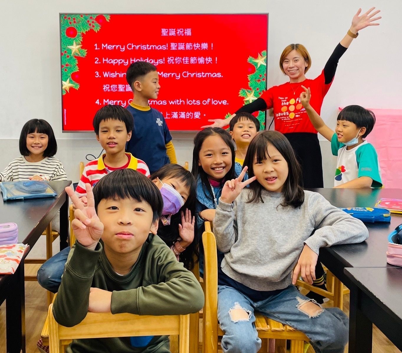 教室裡,孩子們和老師合照。紅色的聖誕節標誌。孩子比劃著和平手勢。