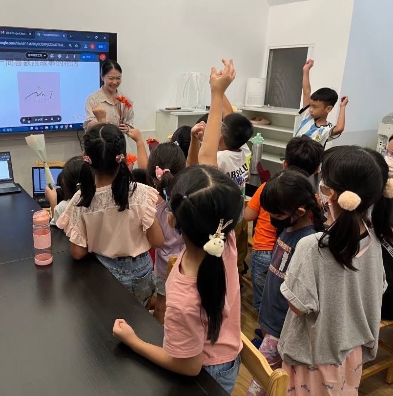 一位老師手持鮮花站在智慧白板前,孩子們在教室裡舉手回答問題。