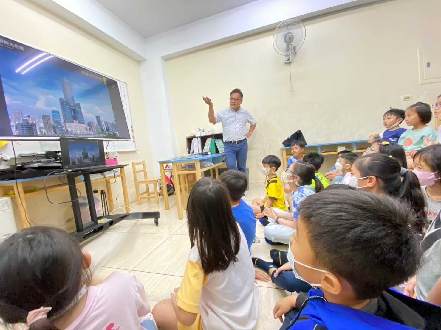 一名男子指著螢幕上顯示的城市景象,向一群坐在地上的孩子講解。