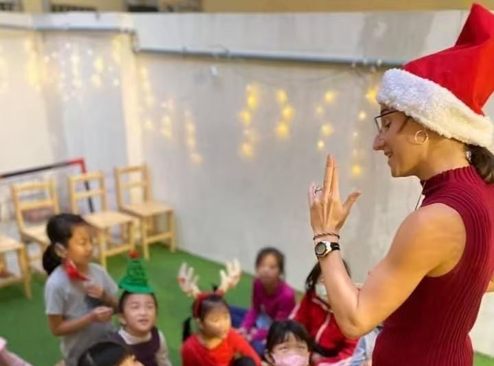 戴著聖誕帽的女子向坐著的孩子們做手勢，背景是一串彩燈。